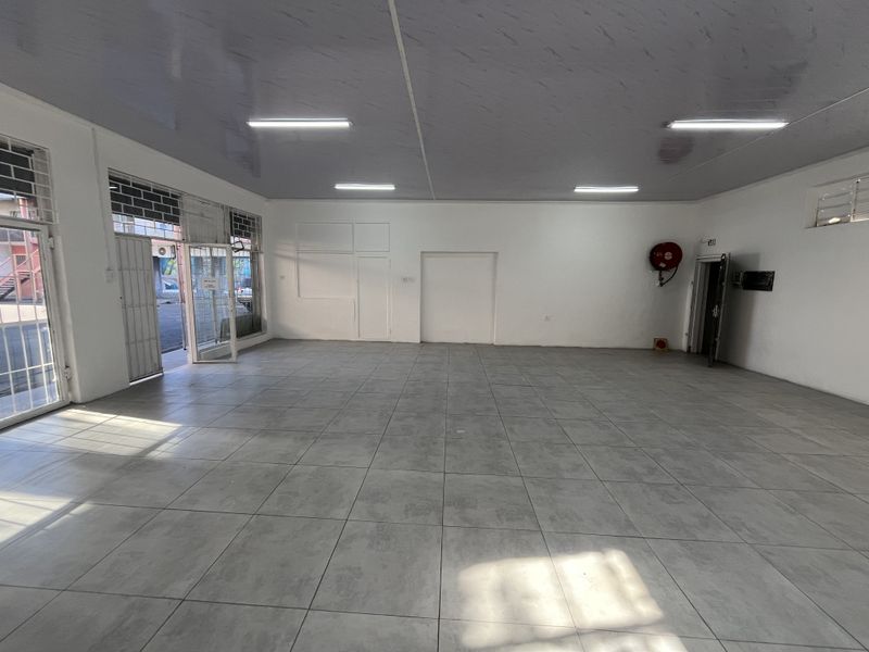 A spacious, empty open-plan retail unit featuring neutral tiled flooring, bright overhead lighting, and secure glass entrance doors.