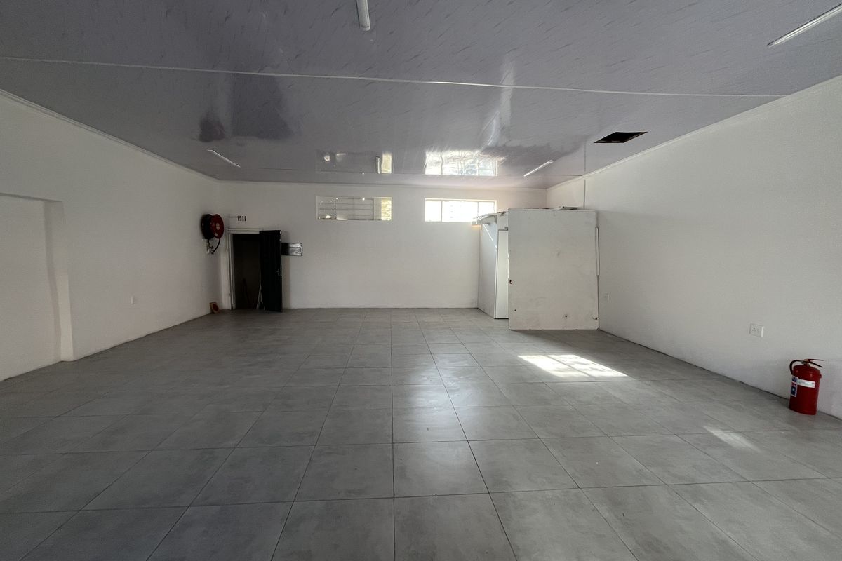 A spacious, empty retail unit featuring modern grey tiled flooring, a clean suspended ceiling, and an integrated cold storage unit.
