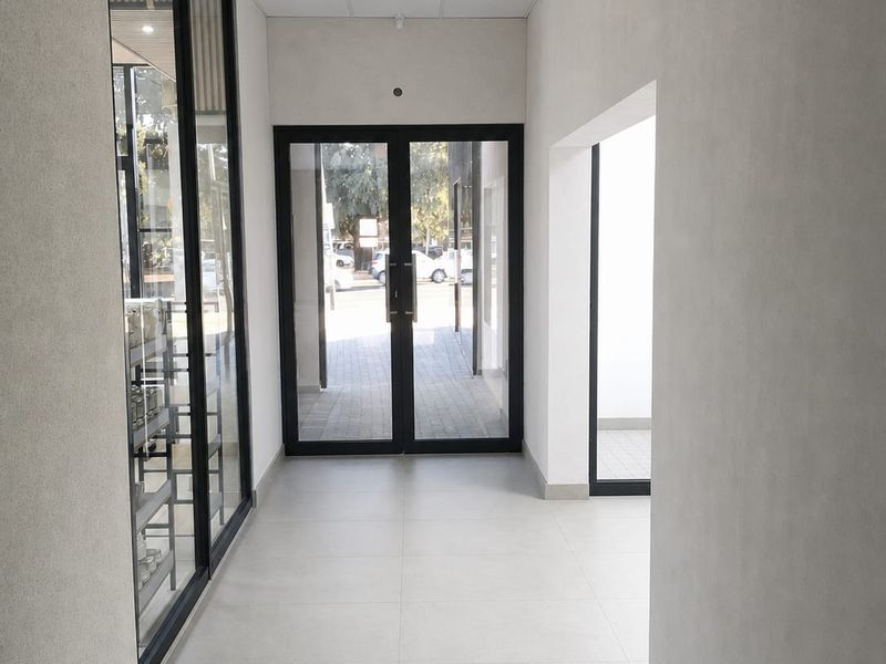 A clean, modern commercial hallway featuring neutral tiled flooring, a suspended ceiling, and large glass entrance doors providing natural light.