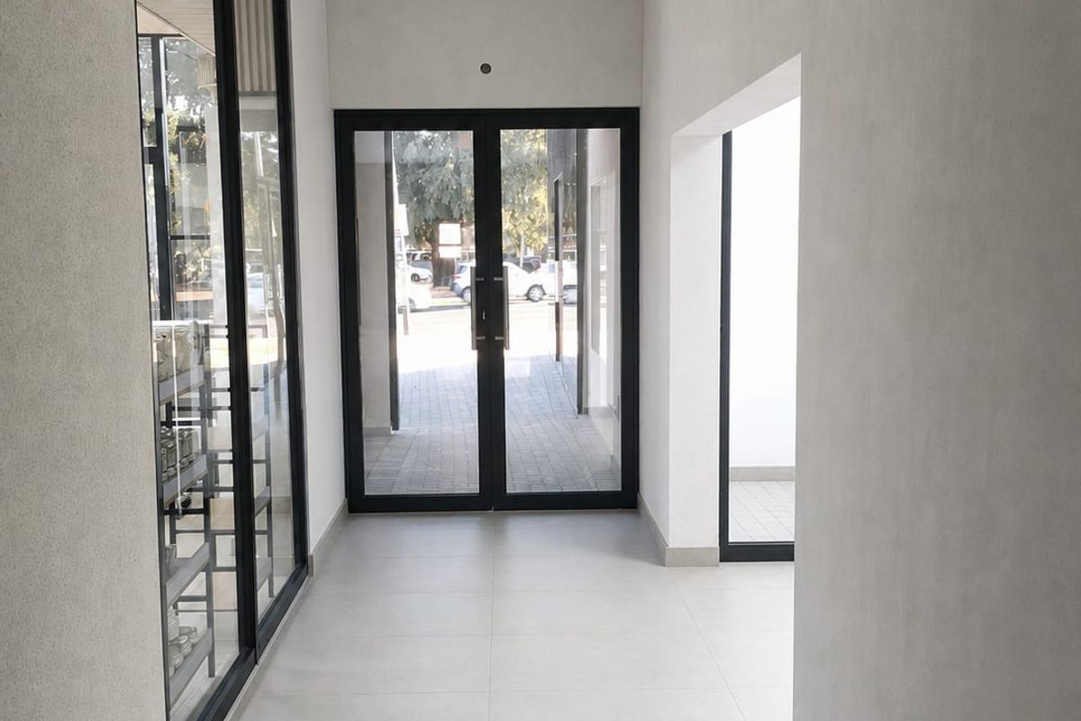 A clean, modern commercial hallway featuring neutral tiled flooring, a suspended ceiling, and large glass entrance doors providing natural light.