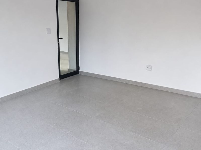 A clean, vacant commercial office space featuring neutral grey floor tiles, white walls, and a modern glass-panelled door.