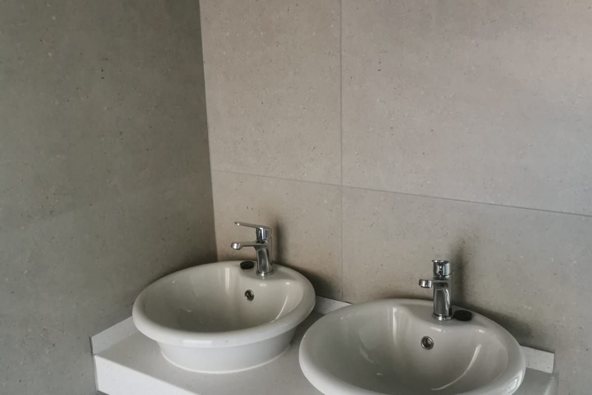 A modern bathroom featuring a double vanity with ceramic basins, chrome faucets, and a stone countertop set against large format neutral wall tiles.