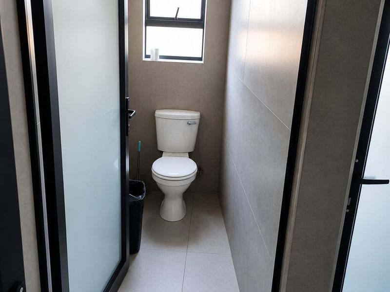 A modern, minimalist guest bathroom featuring neutral floor-to-ceiling tiling, a sleek toilet, and a frosted glass door for privacy.