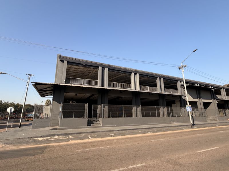 A modern commercial building exterior featuring a multi-level open-plan structure with dark grey facade, balcony railings, and perimeter fencing along the street.