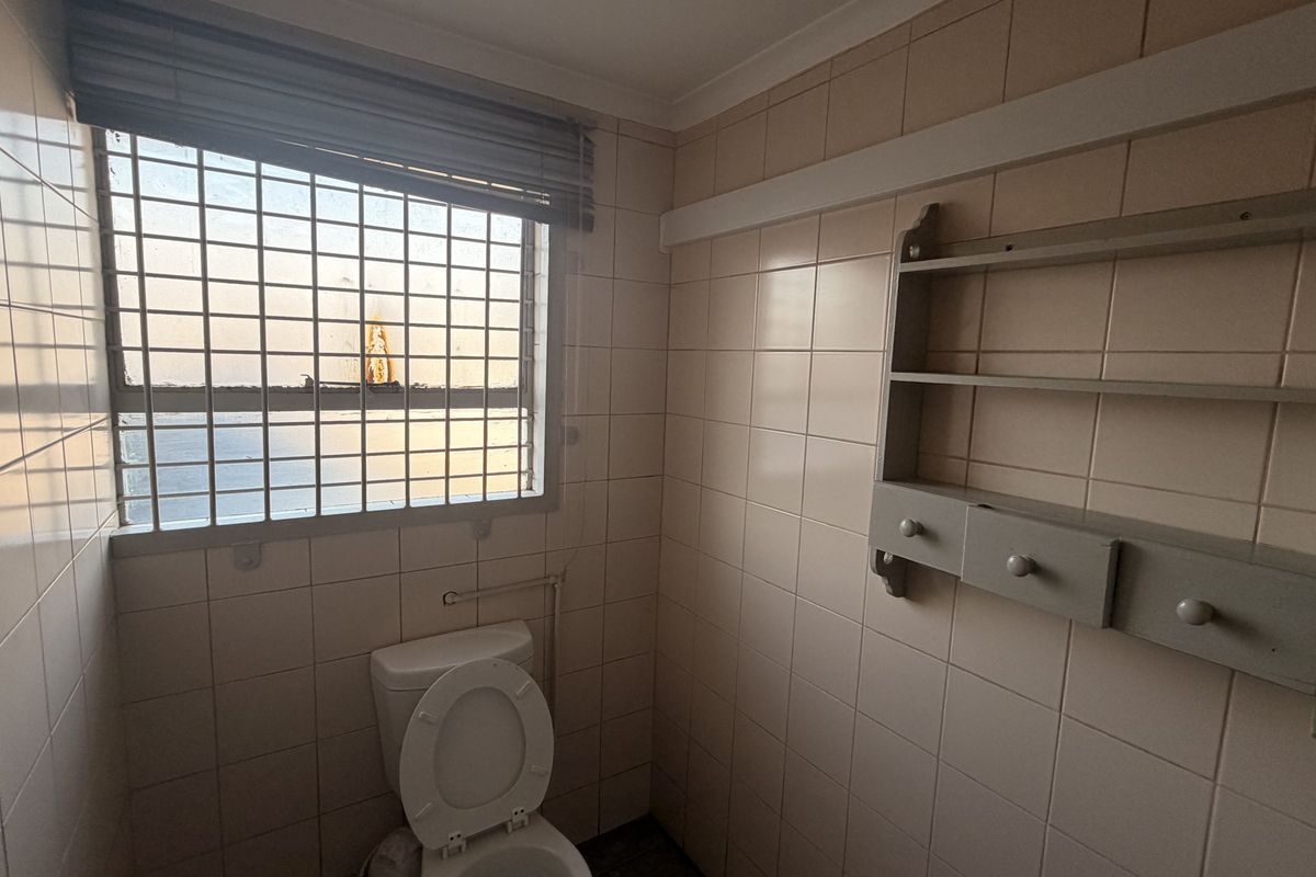A compact bathroom featuring full-height ceramic wall tiling, a standard toilet, and a window fitted with security bars and a blind.