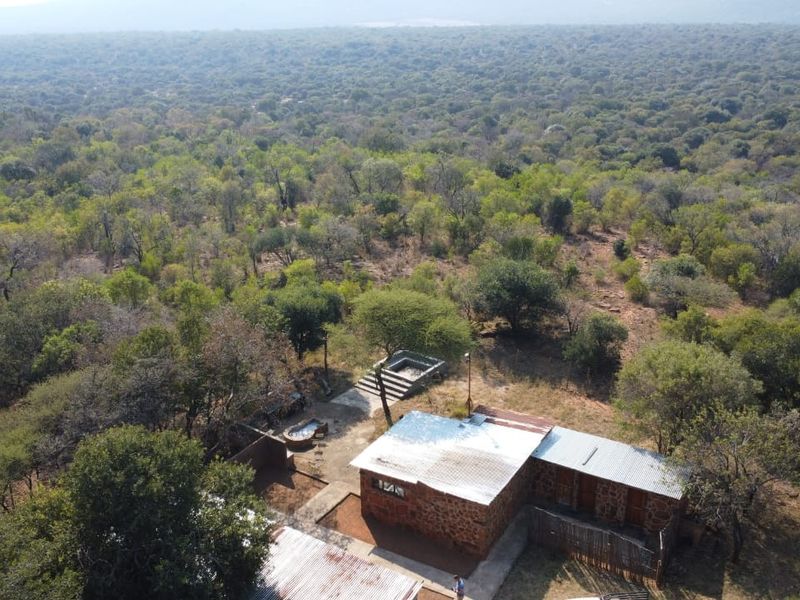Aerial view of a rustic stone farmhouse with a corrugated metal roof, situated within a vast, wooded rural landscape.