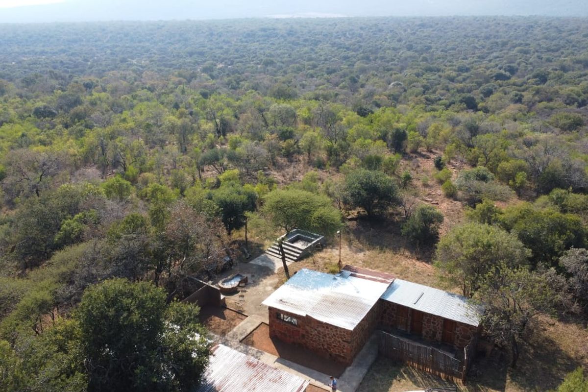 Aerial view of a rustic stone farmhouse with a corrugated metal roof, situated within a vast, wooded rural landscape.