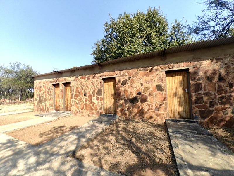 Exterior view of a rustic stone-clad farm building featuring multiple timber entrance doors and concrete pathways under a corrugated metal roof.