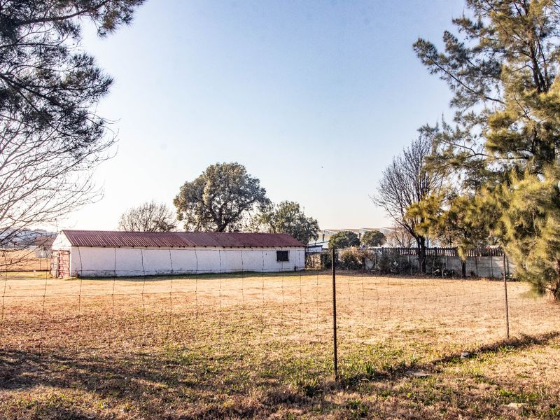 A view of a large open plot featuring a long, single-story outbuilding with a corrugated metal roof and perimeter wire fencing.