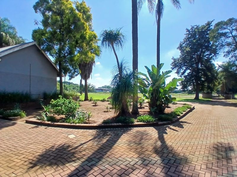 A well-maintained garden area featuring mature trees, landscaped beds, and a brick-paved driveway leading to the office building.