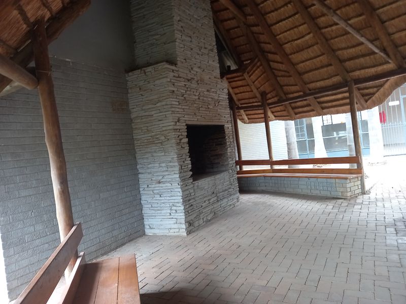 A covered outdoor patio area featuring a stone fireplace, thatched roof, and built-in wooden bench seating on brick paving.
