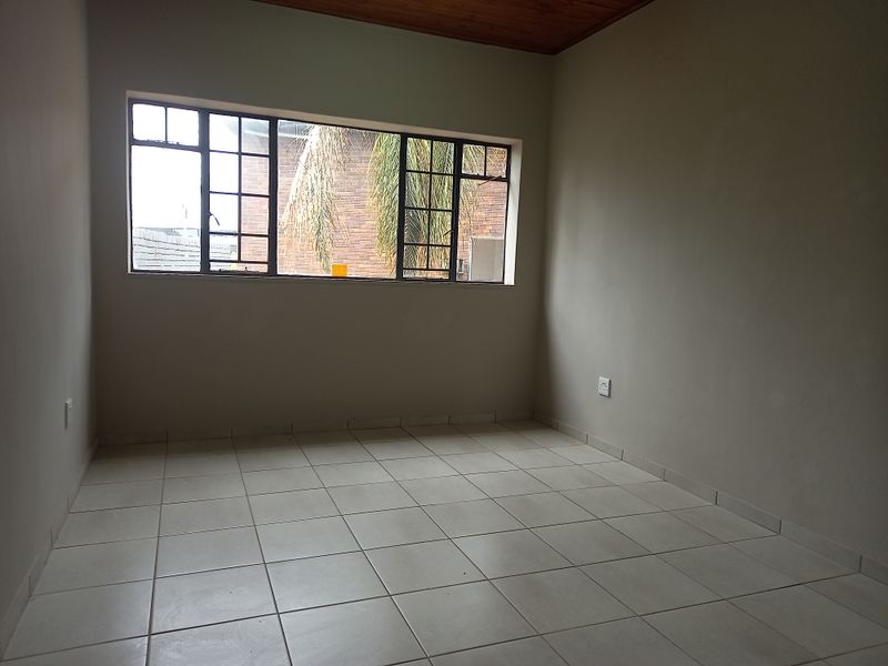 A clean, empty office space featuring neutral tiled flooring, a warm timber ceiling, and a large steel-framed window providing natural light.