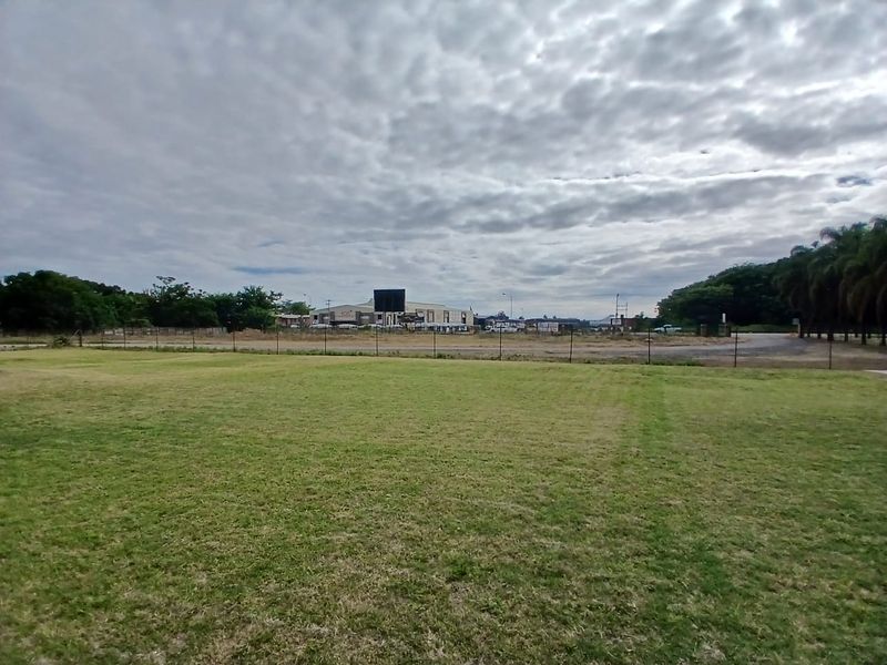 A wide view of a large, open grassed area enclosed by perimeter fencing, with commercial buildings visible in the distance.