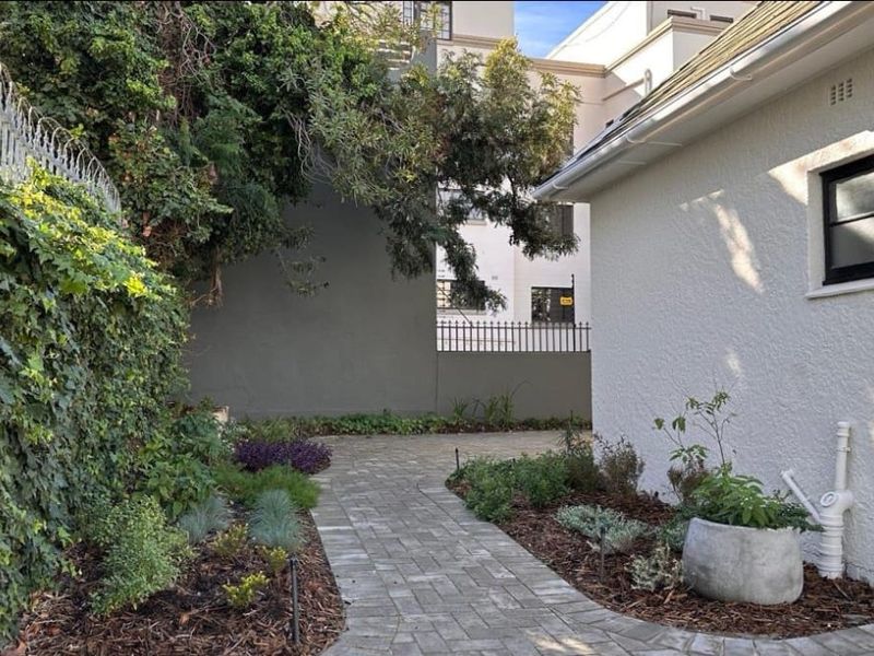 A well-maintained rear exterior area featuring a paved walkway, landscaped garden beds, and a clean white rendered building facade.