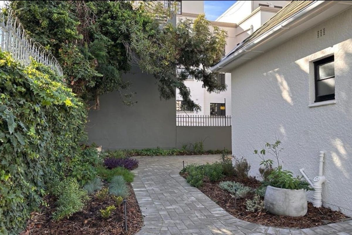 A well-maintained rear exterior area featuring a paved walkway, landscaped garden beds, and a clean white rendered building facade.