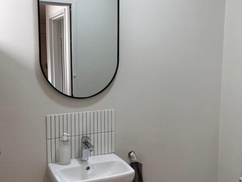 A clean and modern office restroom featuring a wall-mounted basin with chrome fixtures, vertical tile splashback, and a contemporary oval mirror.