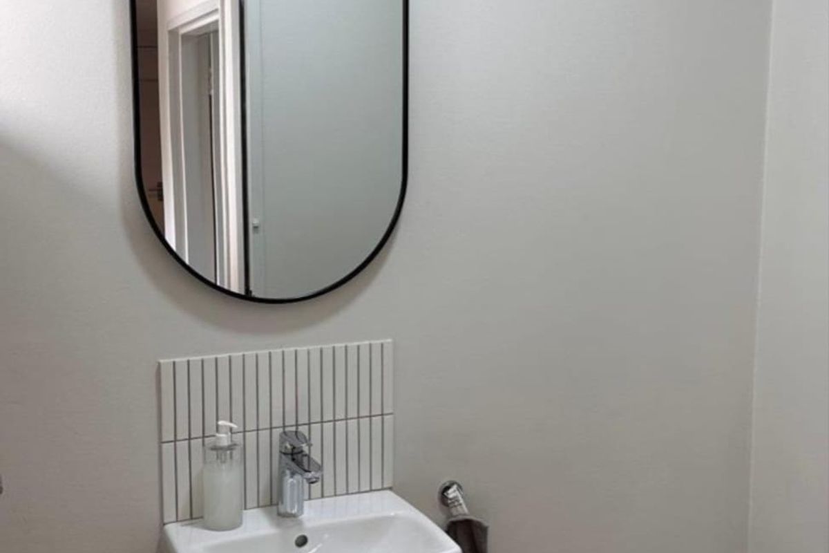 A clean and modern office restroom featuring a wall-mounted basin with chrome fixtures, vertical tile splashback, and a contemporary oval mirror.