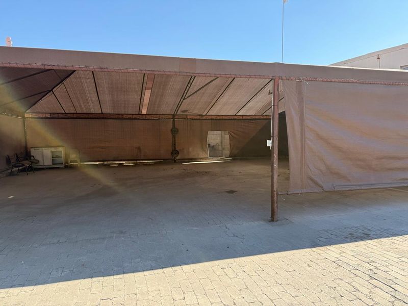 A large outdoor covered area featuring a metal frame structure with canvas shade covering and paved flooring, suitable for commercial storage or utility use.
