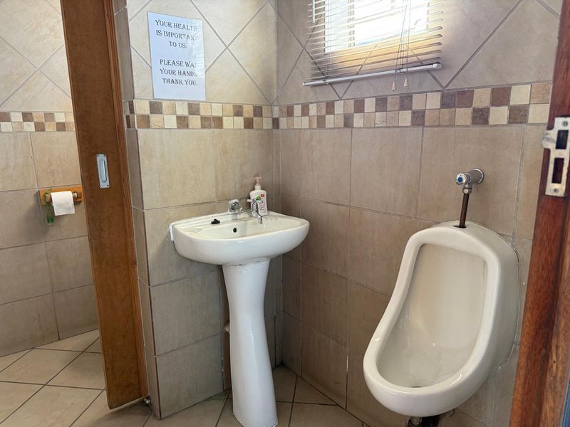 A functional commercial restroom featuring a pedestal washbasin, wall-mounted urinal, and neutral ceramic tiling throughout.