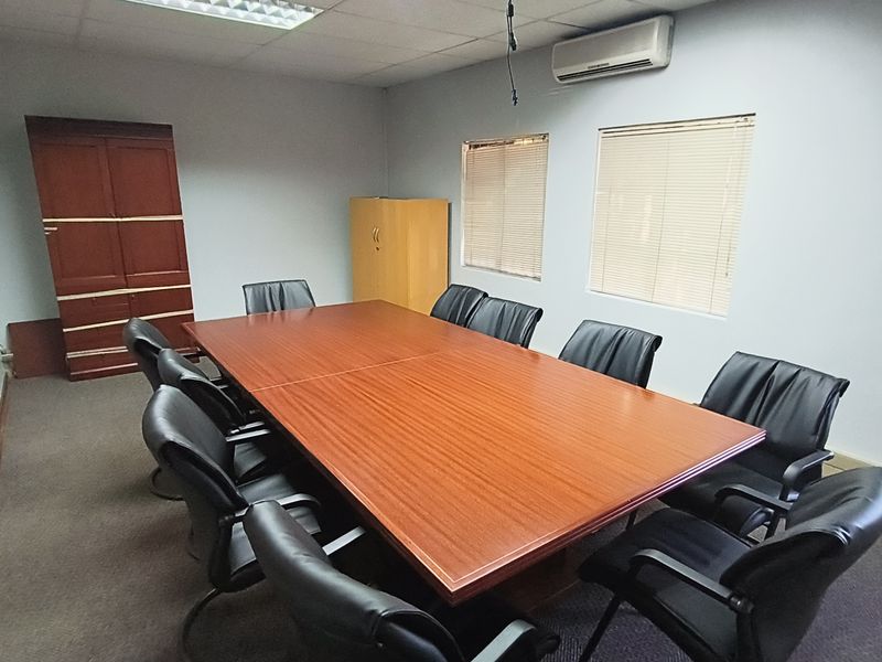 A functional office meeting room featuring a large boardroom table, carpeted flooring, suspended ceiling, and wall-mounted air conditioning unit.