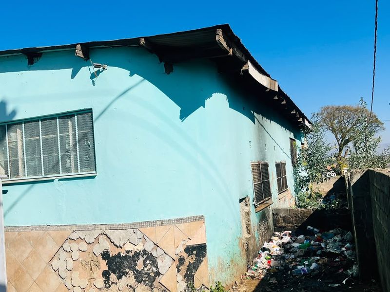 Rear exterior view of the property showing a painted facade with security-barred windows, damaged wall tiling, and significant debris accumulation along the perimeter wall.