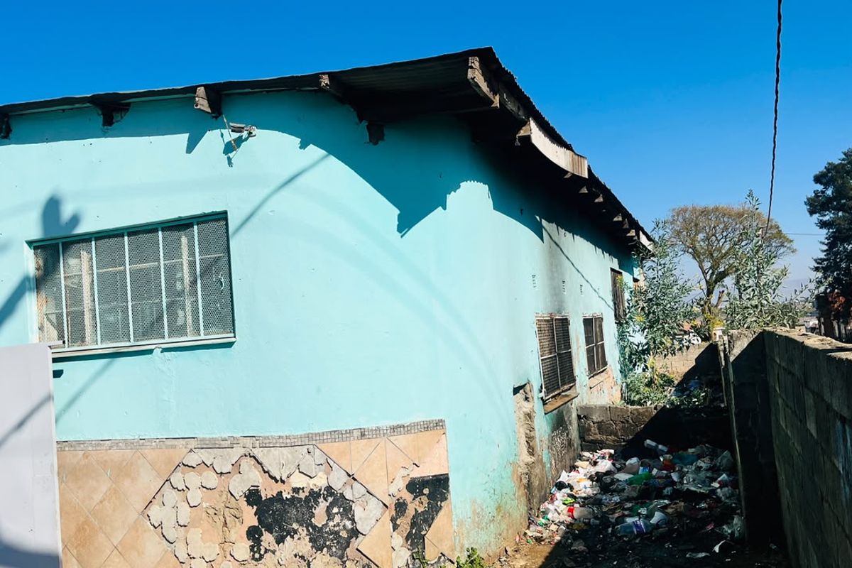 Rear exterior view of the property showing a painted facade with security-barred windows, damaged wall tiling, and significant debris accumulation along the perimeter wall.
