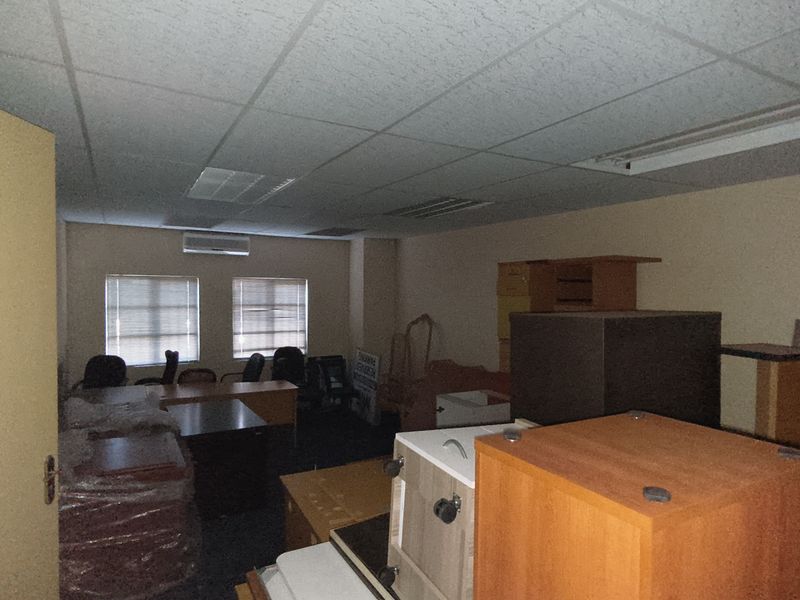 A cluttered office space featuring a suspended ceiling, recessed lighting, and a wall-mounted air conditioning unit, currently used for storage.