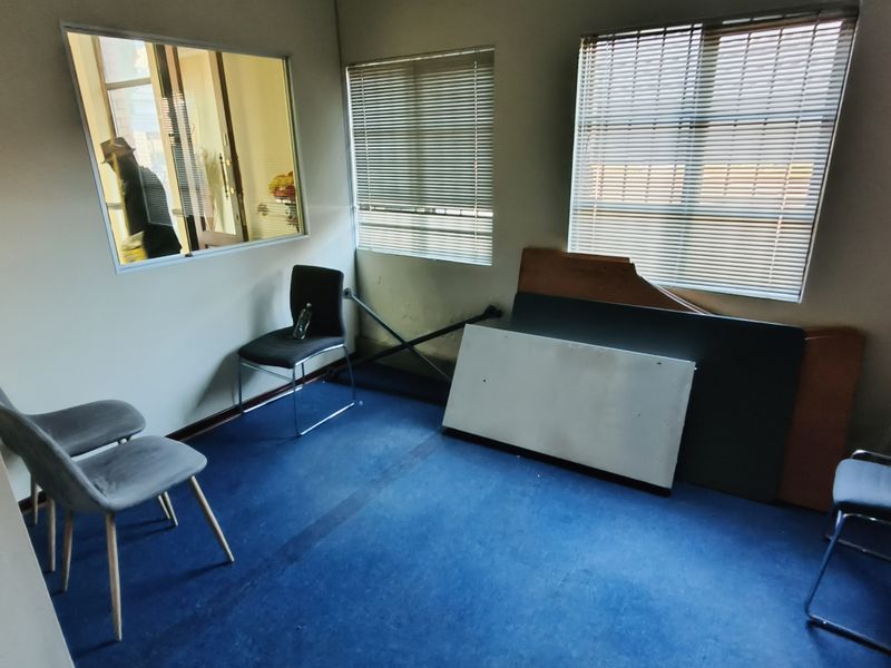 A small office space featuring blue carpet flooring, internal window partition, and venetian blinds on the exterior windows.
