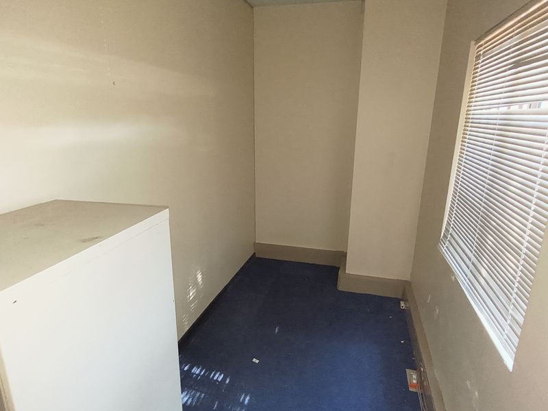 A small, empty office space featuring blue carpet flooring and a window fitted with venetian blinds.