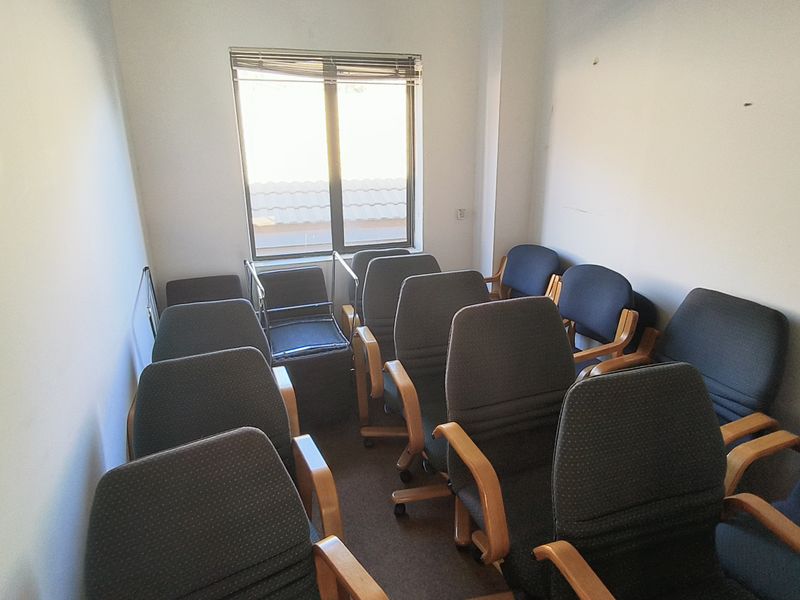 A small office space featuring carpet flooring and a window with blinds, currently set up with multiple chairs.