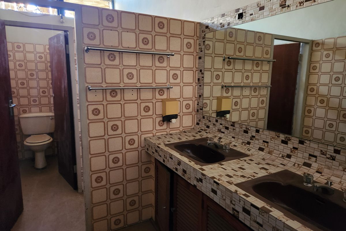A dated industrial bathroom featuring a double vanity with tiled countertops, wall-mounted mirror, and a separate toilet area with vintage wall tiles.