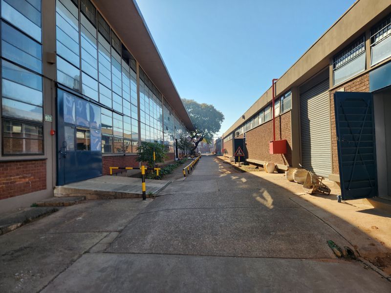 An industrial exterior corridor featuring concrete paving, large steel-framed windows, and roller shutter access points between two warehouse structures.