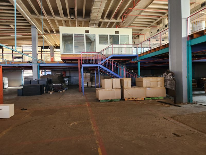 Spacious industrial warehouse interior featuring a mezzanine level with an elevated office pod, steel staircase, and exposed concrete structural elements.