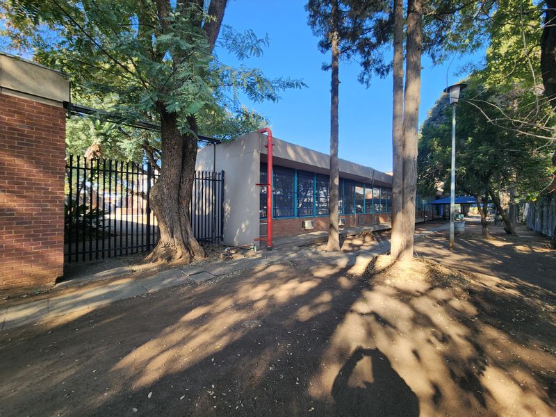 Exterior view of an industrial building featuring brick masonry, metal security fencing, and large windows, set within a landscaped area with mature trees.