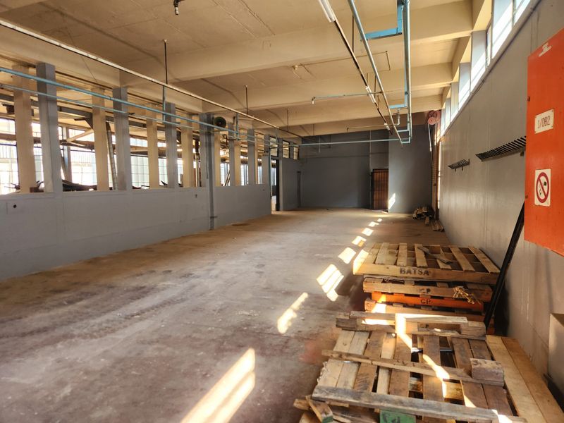 Spacious industrial utility area featuring exposed concrete ceilings, overhead piping, and large steel-framed windows, suitable for workshop or storage use.