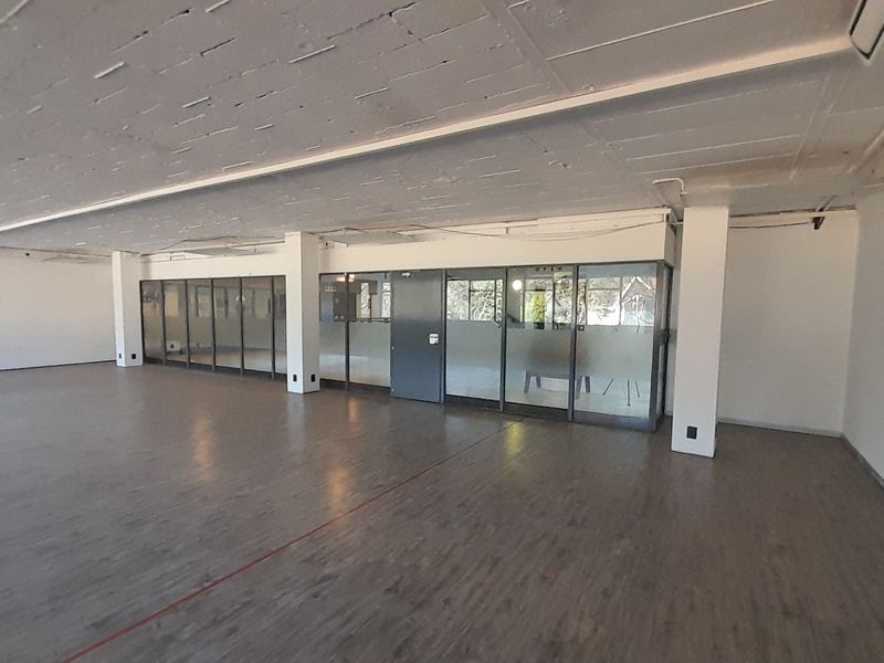Spacious open-plan office area featuring exposed concrete ceilings, laminate flooring, and glass-partitioned meeting rooms, offering a versatile workspace layout.