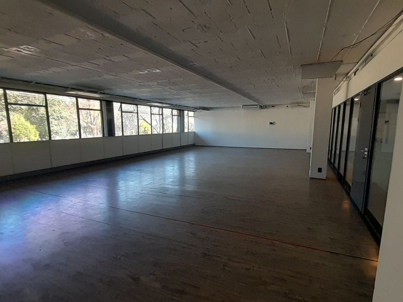 A spacious, open-plan office area featuring large windows, exposed concrete ceilings, and modern glass partitions, offering a bright and versatile workspace.