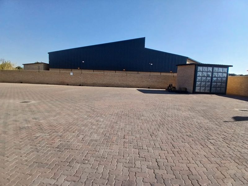 A spacious paved industrial yard featuring a brick perimeter wall and a large metal-clad warehouse structure.
