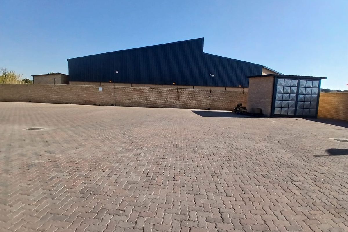 A spacious paved industrial yard featuring a brick perimeter wall and a large metal-clad warehouse structure.