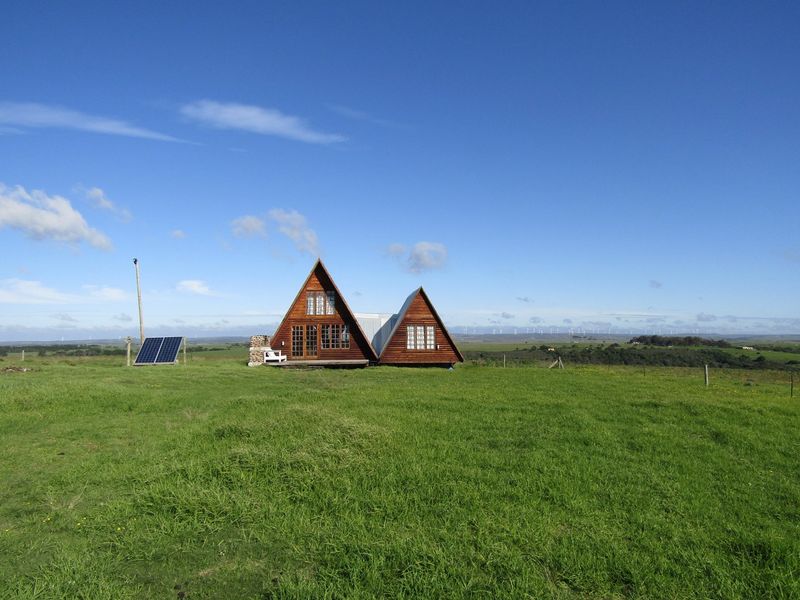 A charming A-frame timber farmhouse set in a vast green field, featuring solar panels and a stone chimney.