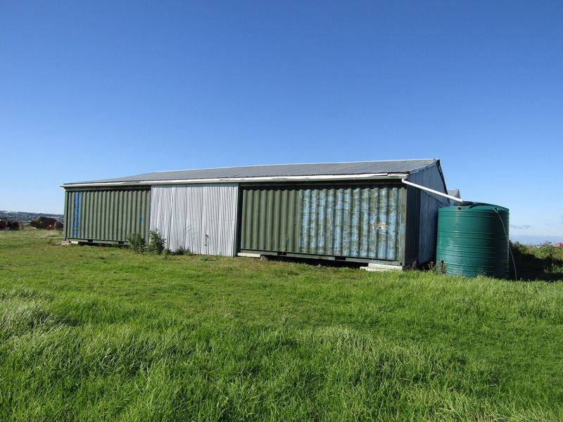 A functional farm outbuilding constructed from shipping containers, featuring a corrugated metal roof and an attached rainwater collection tank.