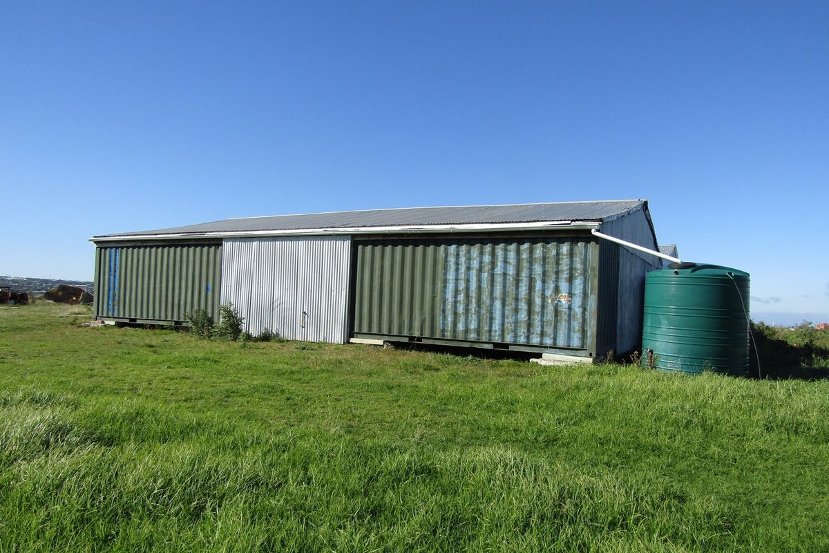A functional farm outbuilding constructed from shipping containers, featuring a corrugated metal roof and an attached rainwater collection tank.