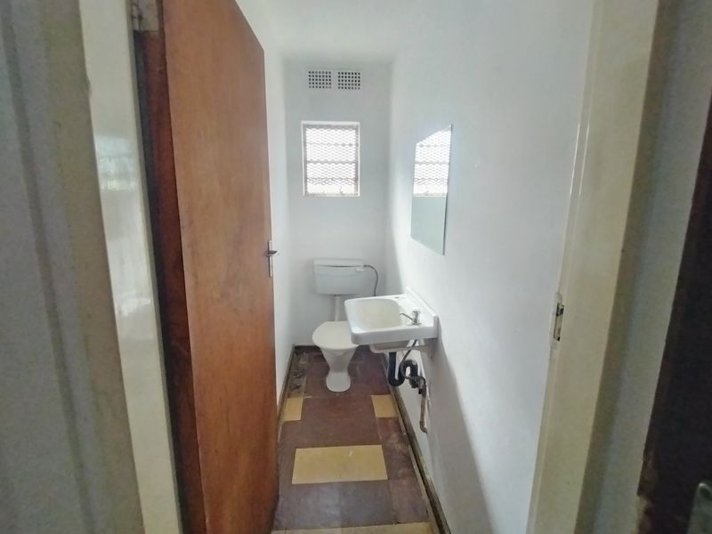 A compact, functional bathroom featuring a wall-mounted basin, toilet, and window with security bars, requiring cosmetic updates to the flooring and finishes.