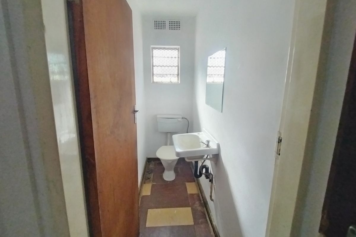 A compact, functional bathroom featuring a wall-mounted basin, toilet, and window with security bars, requiring cosmetic updates to the flooring and finishes.
