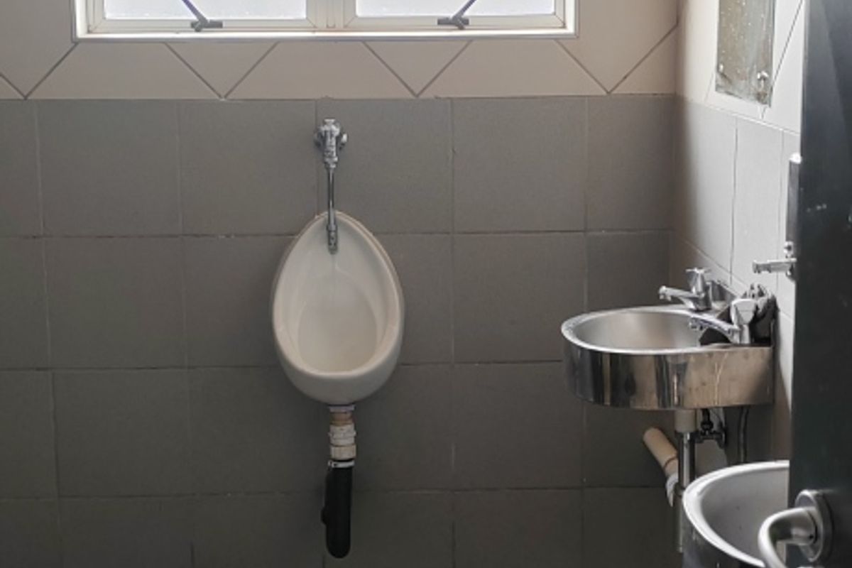 Functional industrial bathroom featuring a wall-mounted urinal, stainless steel basin, tiled finishes, and natural light from a casement window.