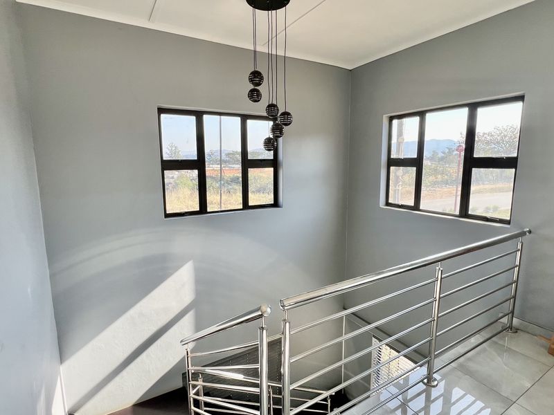 A bright, modern hallway featuring a stainless steel balustrade, tiled flooring, and contemporary pendant lighting with large windows providing natural light.