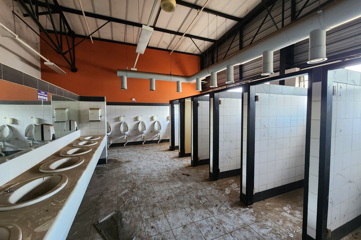 Industrial style communal bathroom featuring multiple urinals, vanity basins, and tiled partitions with exposed ceiling structure, currently requiring significant maintenance and cleaning.