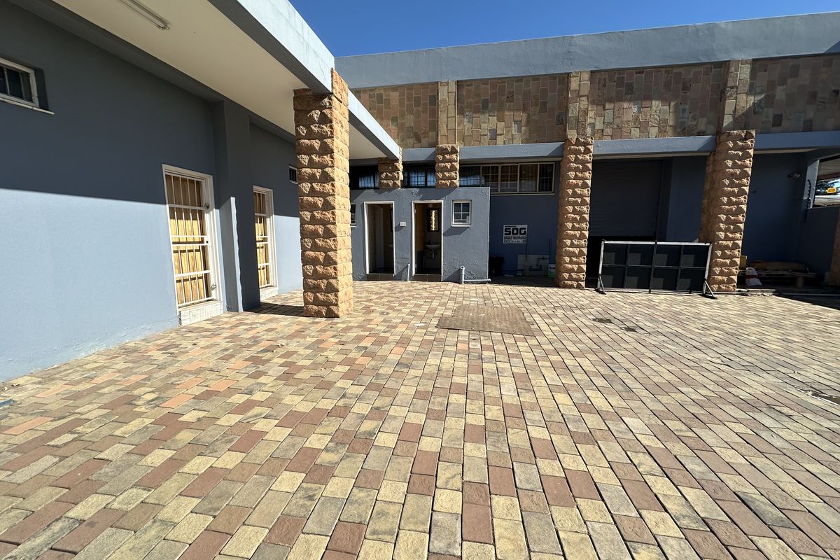 A paved industrial courtyard featuring stone-clad pillars, security gates, and a roller shutter door access point.