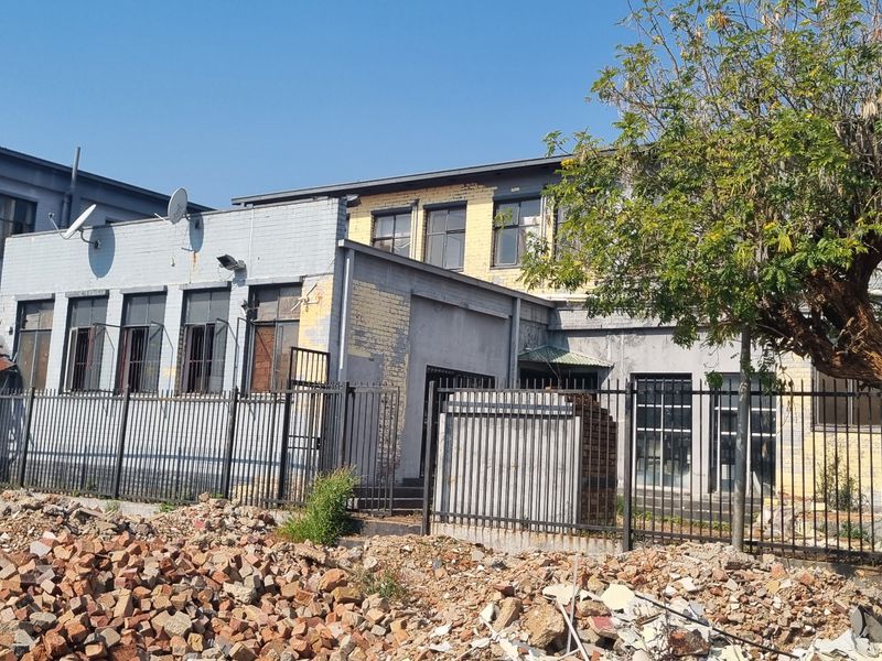 Rear exterior view of an industrial-style building featuring brick masonry, metal security fencing, and multiple windows showing signs of significant wear and deterioration.