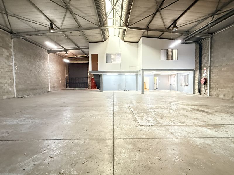 Spacious industrial warehouse featuring high ceilings with exposed steel trusses, concrete flooring, a mezzanine level, and a large roller shutter door.